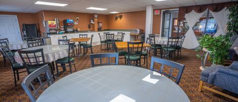 A dining area with round tables and chairs, a kitchen in the background, and a plant on the right.