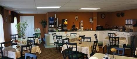 A dining area with tables and chairs, a kitchen area with appliances, and a clock on the wall.