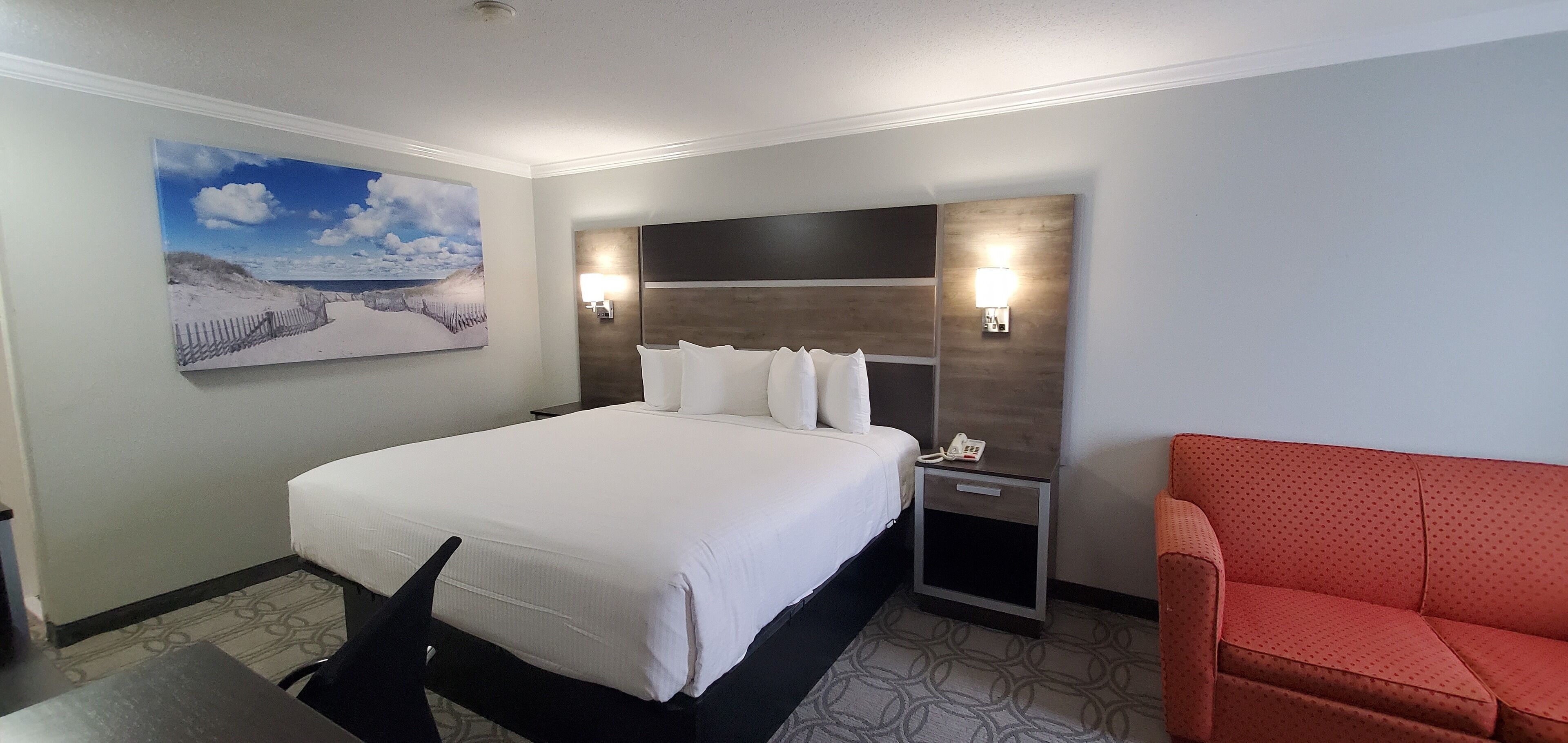 A hotel room with a large bed, a nightstand with a phone, a red chair, and a painting of a beach scene.