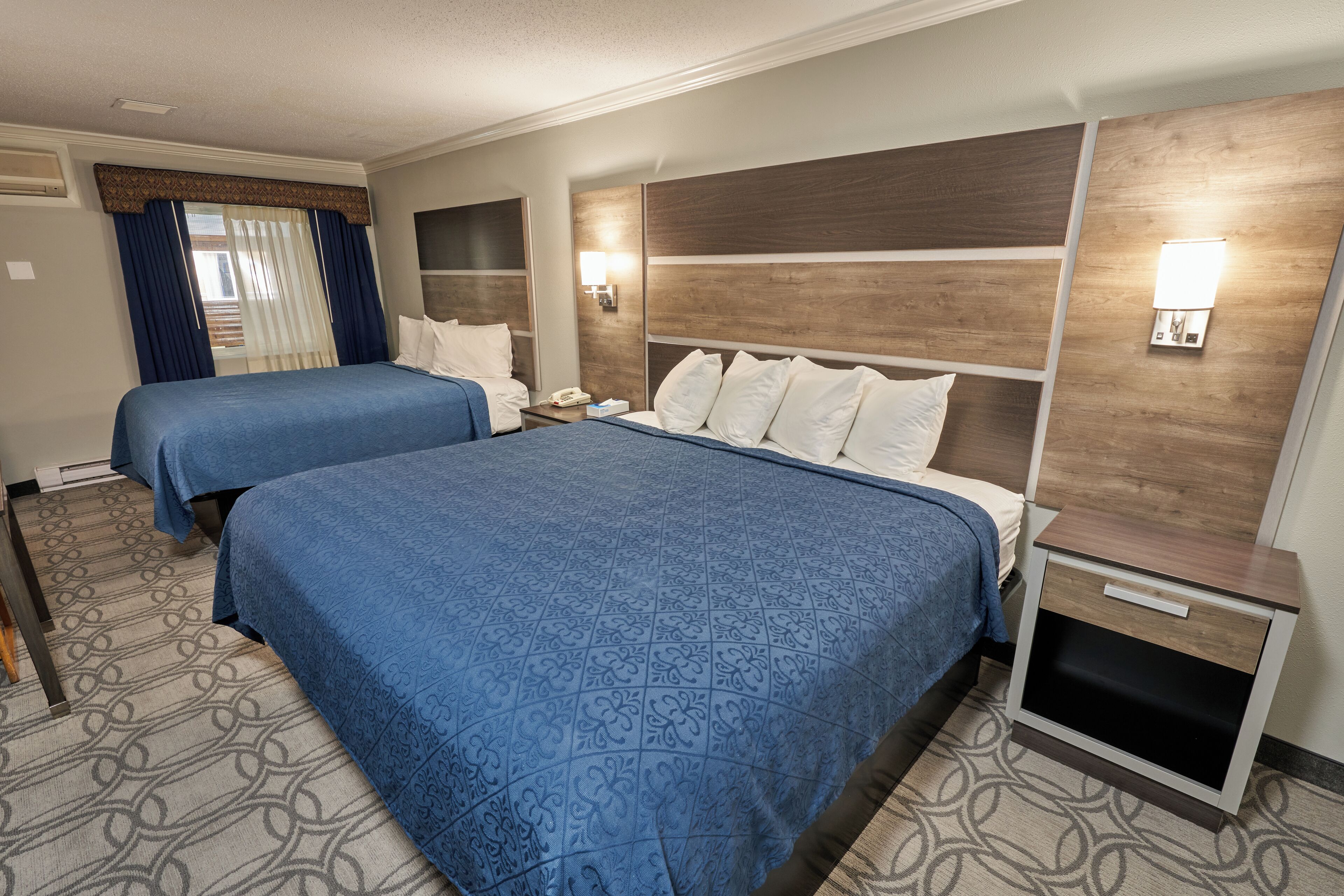 A hotel room with two beds, a wooden headboard, a nightstand with a phone, and a wall-mounted light.