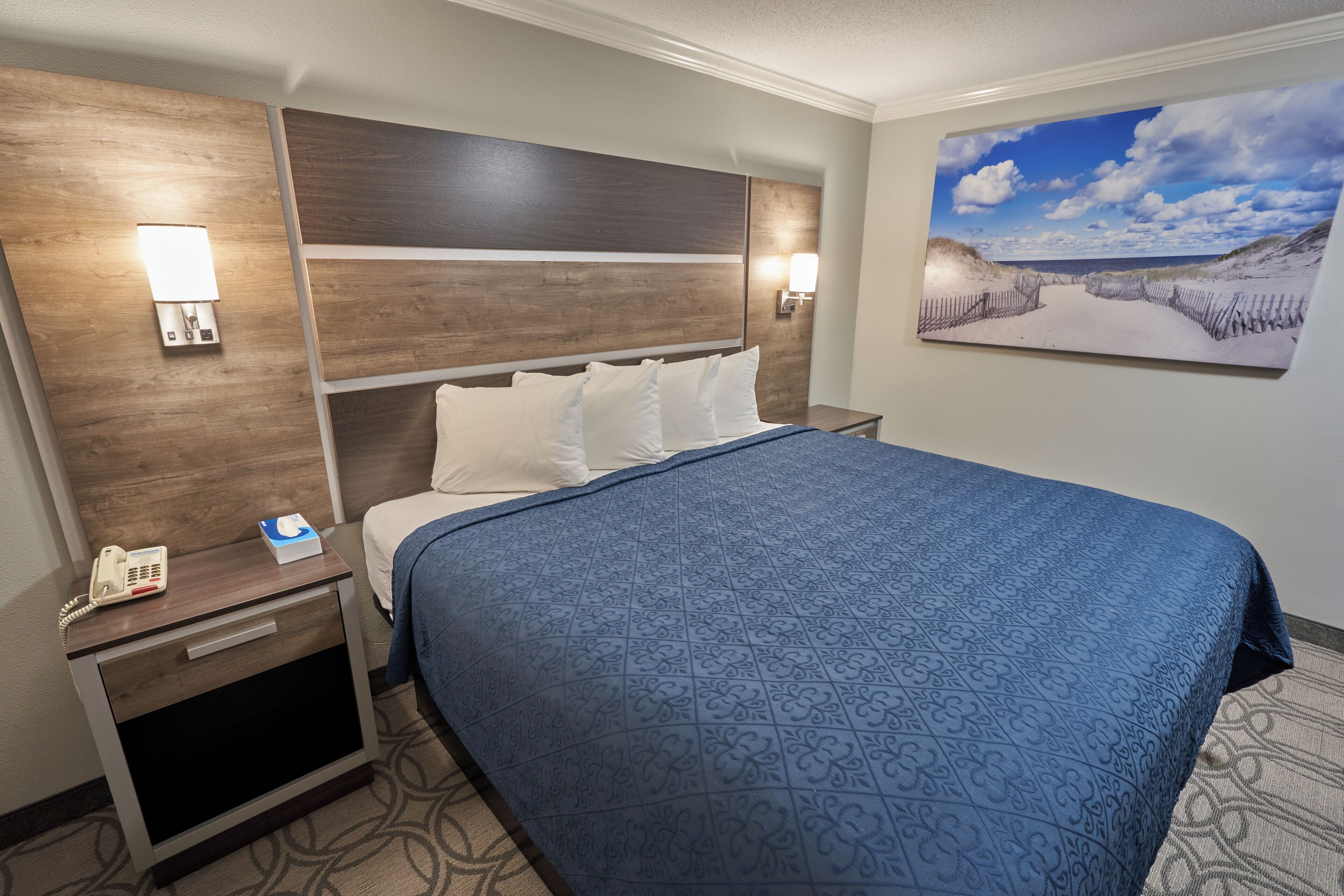 Une chambre d’hôtel avec un grand lit, une tête de lit en bois, une table de chevet avec un téléphone et un tableau représentant une scène de plage.