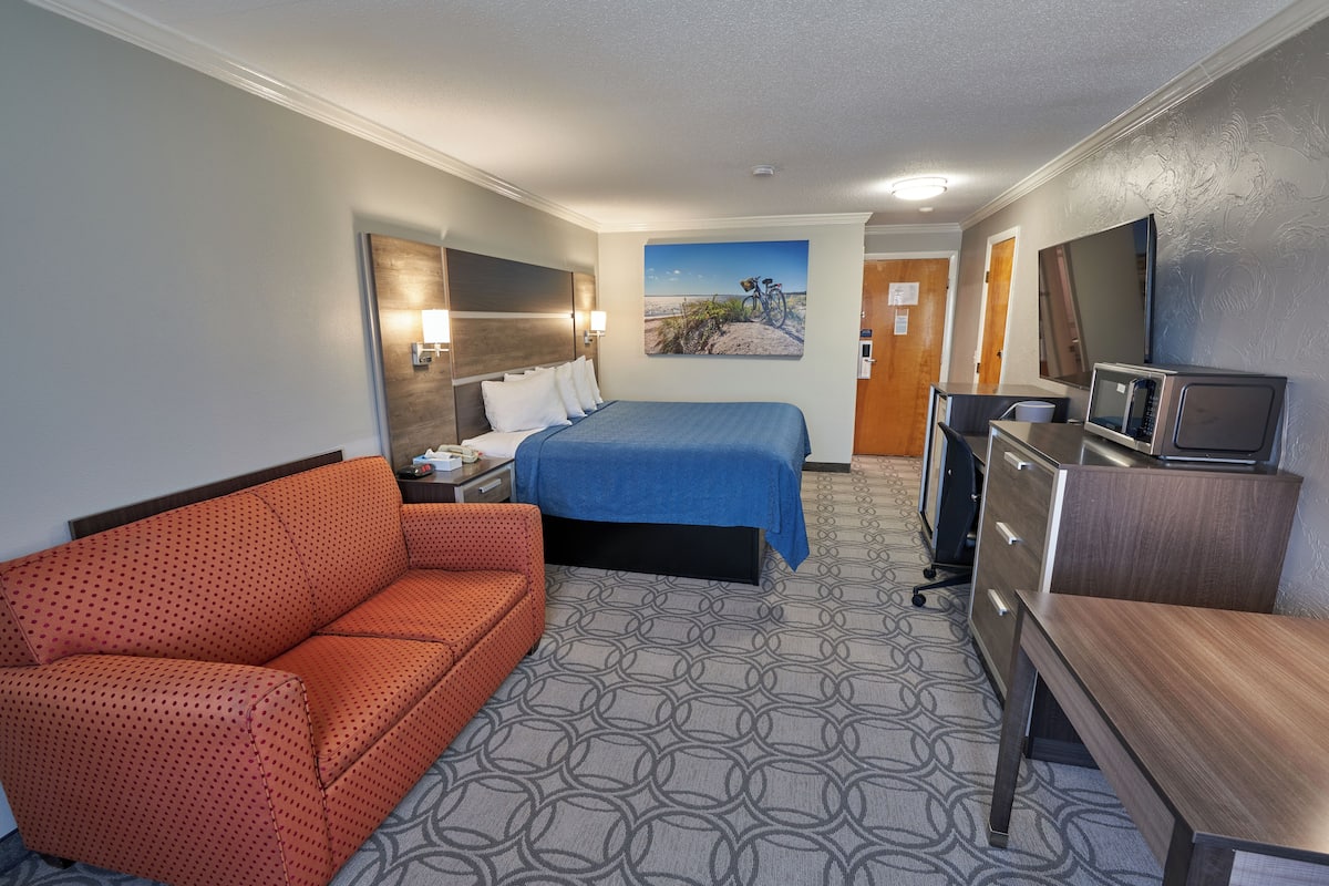 A hotel room with a bed, a sofa, a desk, a television, and a painting on the wall.