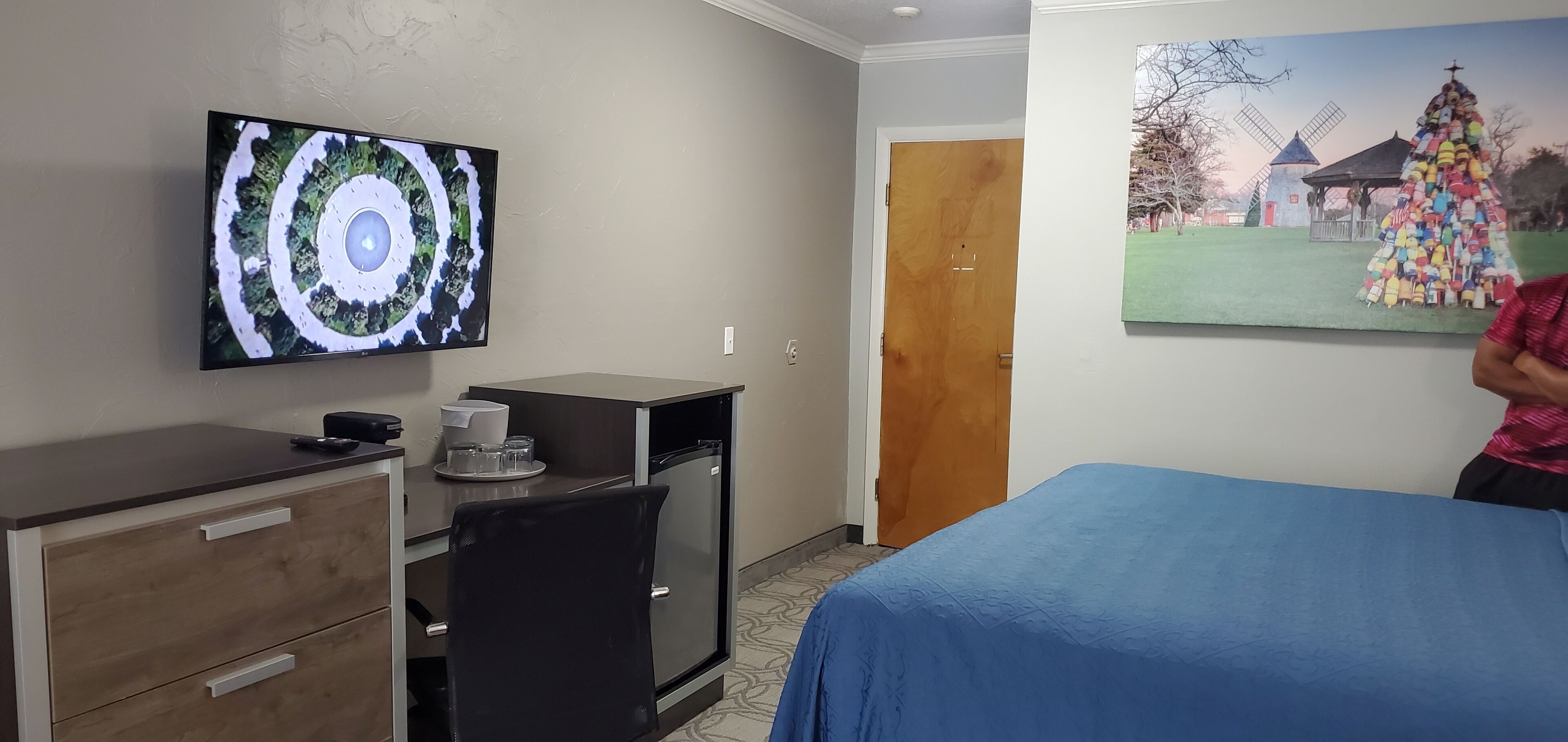 Una habitación de hotel con una cama, un escritorio con un ordenador, un televisor en la pared y un cuadro de un molino.