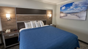 A hotel room with a large bed, a headboard with built-in lighting, a bedside table with a phone and a remote control, and a wall-mounted painting of a beach scene. - Ambassador Inn & Suites (South Yarmouth)