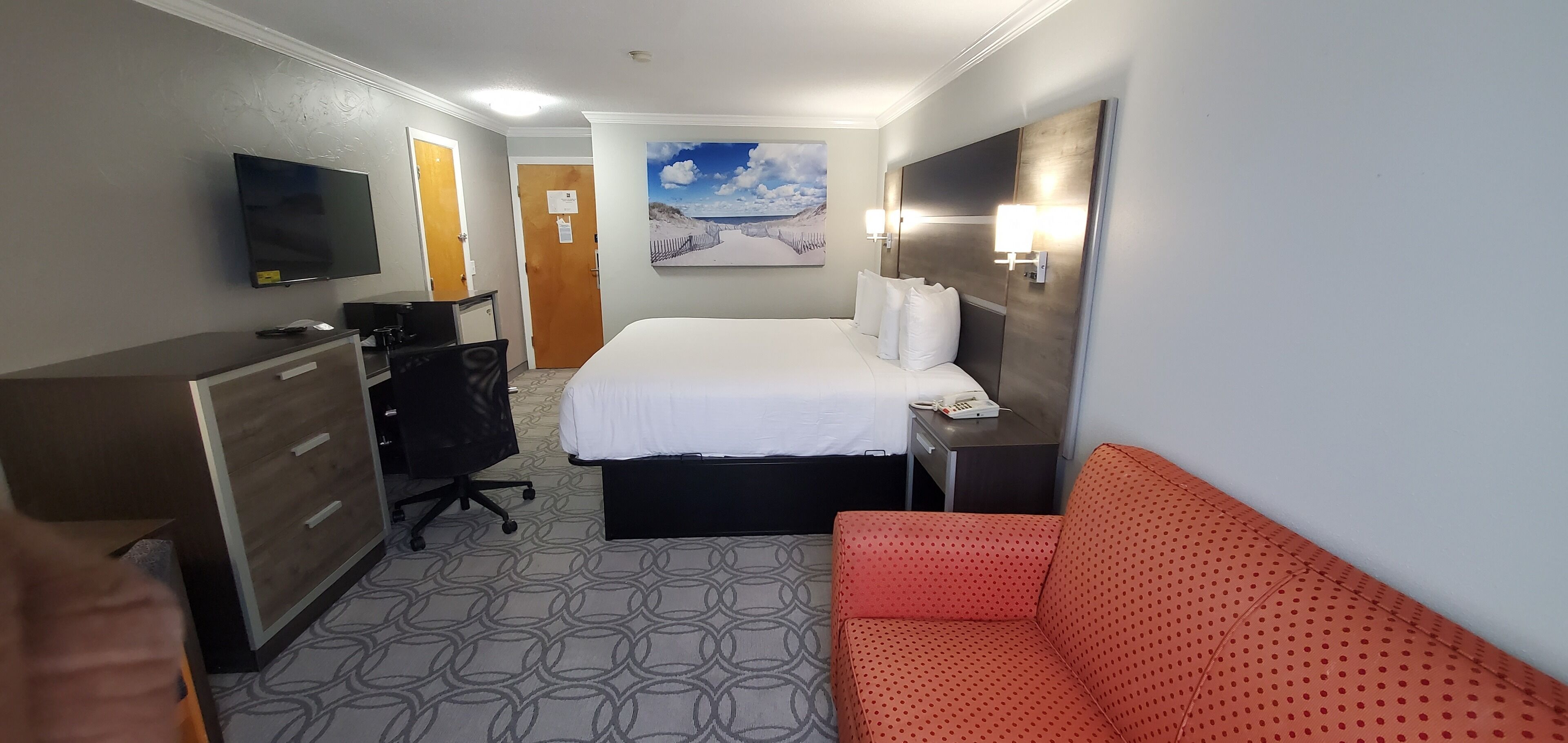 Habitación de hotel con cama, escritorio, silla, televisor, un cuadro y sofá.