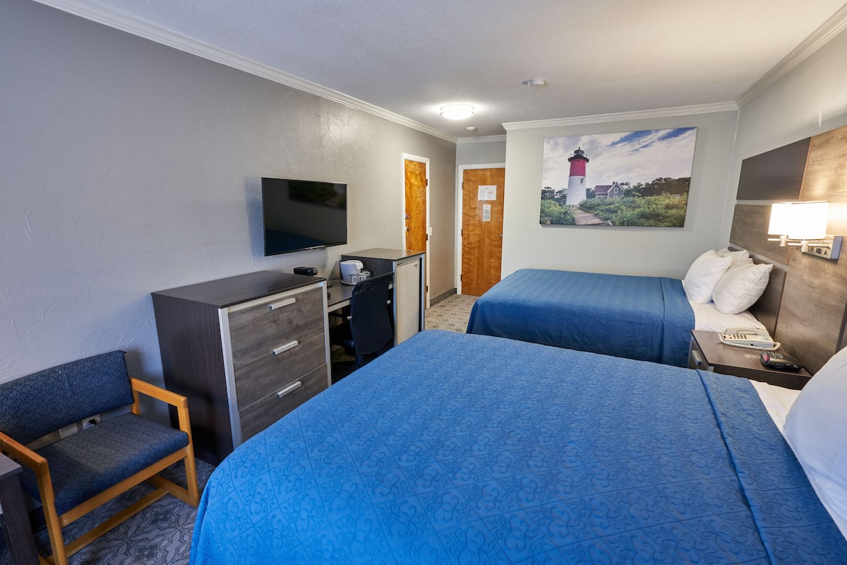 A hotel room with two beds, a desk, a chair, a TV, and a painting of a lighthouse.
