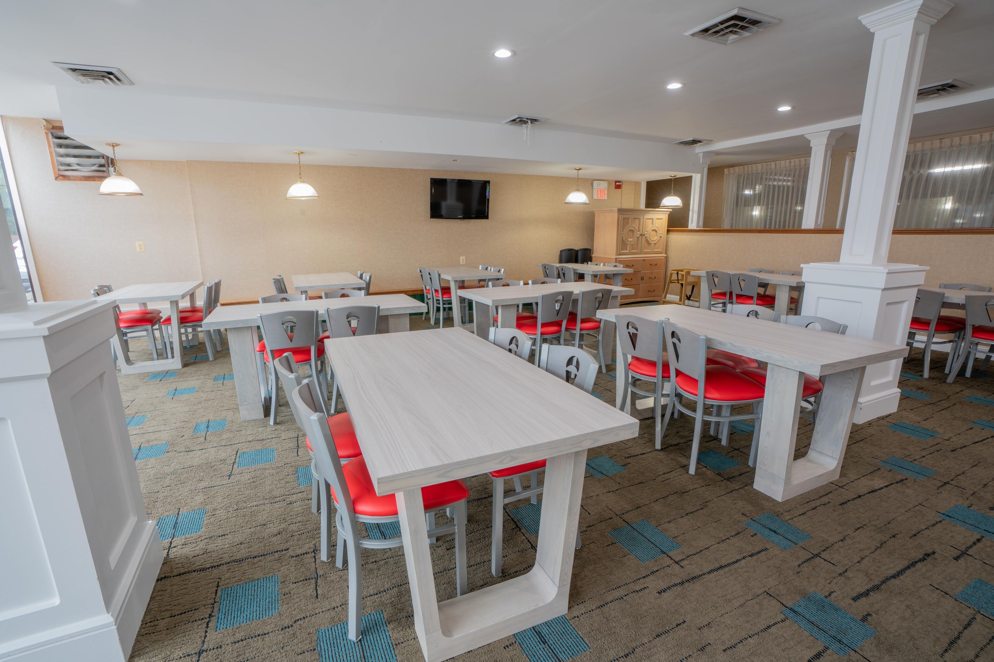 A dining area with multiple tables and chairs, a television mounted on the wall, and patterned carpet.