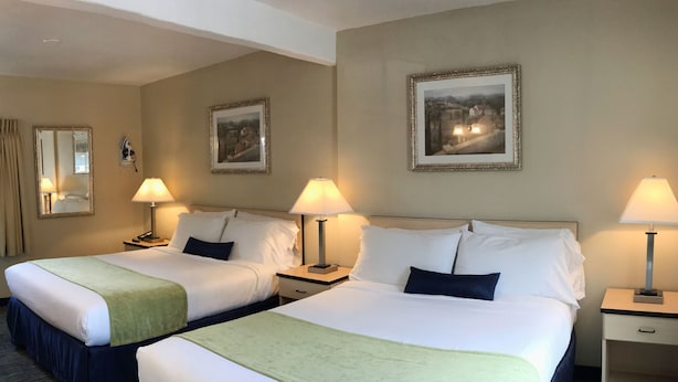 A hotel room with two beds, each with a green throw blanket, white bedding, and matching nightstands with lamps. There are framed pictures on the walls and a mirror above the beds.