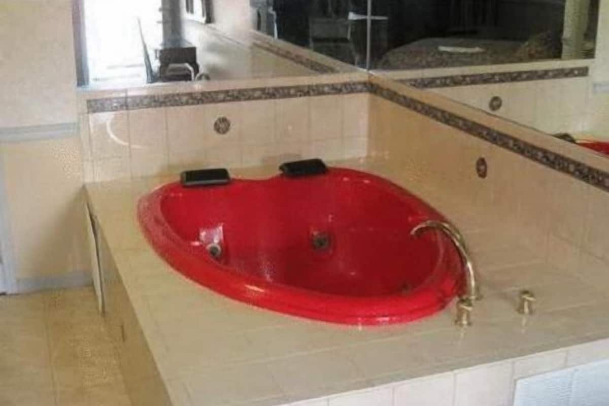 Una bañera roja en un baño con azulejos color crema y un espejo encima.