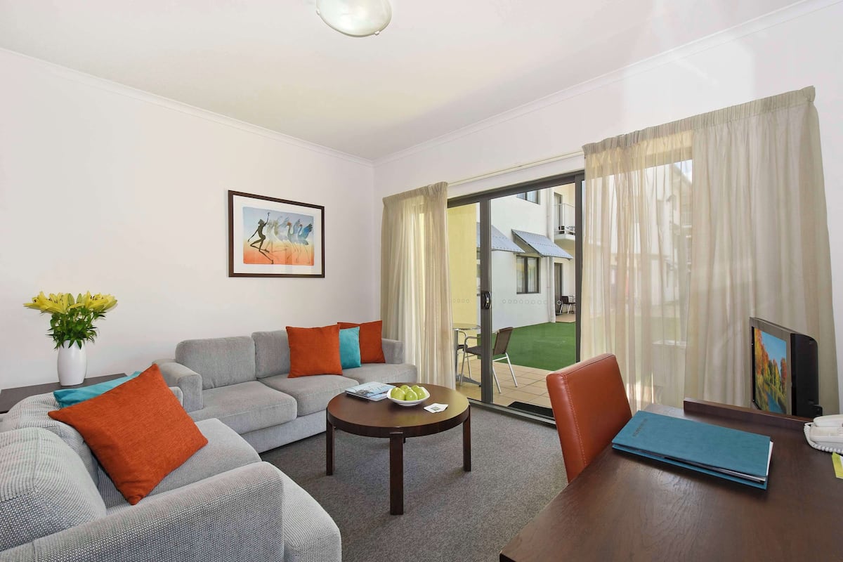 A modern living room with a grey sofa, orange pillows, a coffee table with a bowl of fruit, and a view of a green lawn through a sliding glass door.