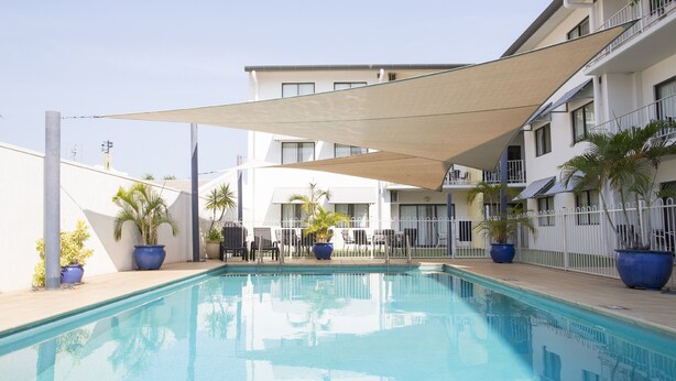 En swimmingpool med liggestole og plantekasser, omgivet af en hvid bygning med altaner.