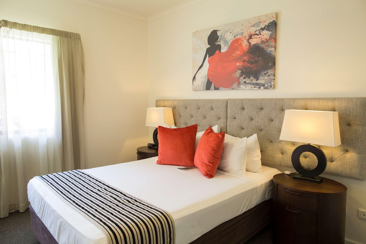 A hotel room with a bed, two bedside lamps, a headboard, a nightstand, and a painting on the wall.
