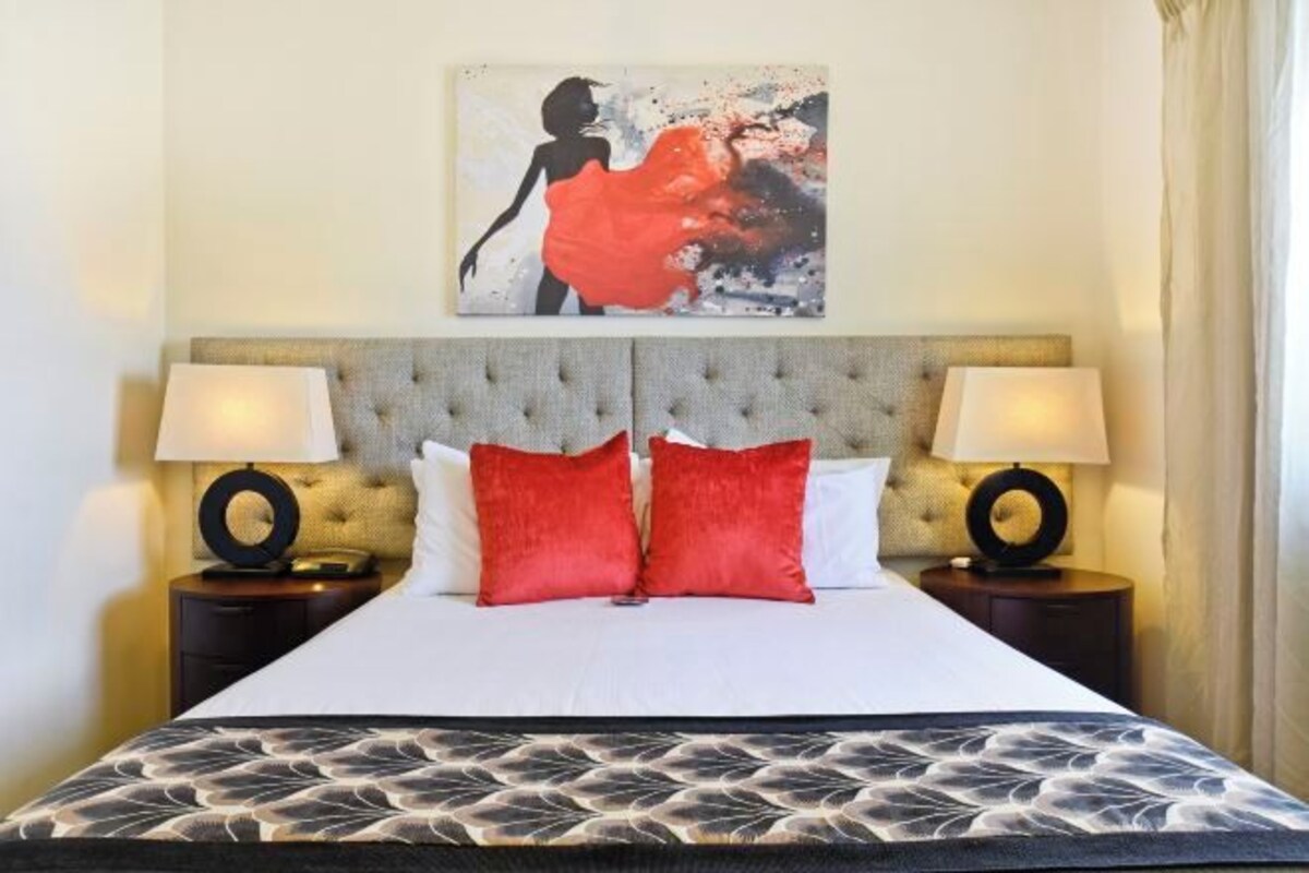 A hotel room with a bed, two bedside tables with lamps, a headboard, and a painting above the bed.