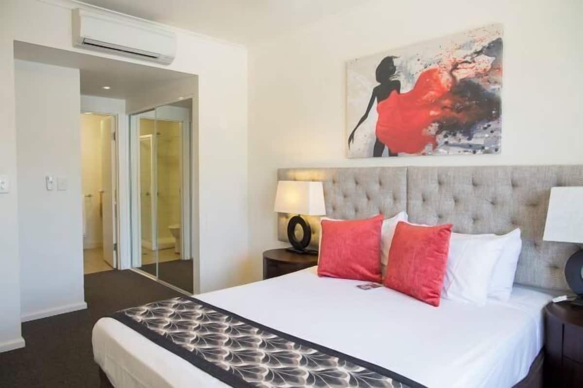 A hotel room with a large bed, a headboard, a painting, a nightstand, and an air conditioning unit.