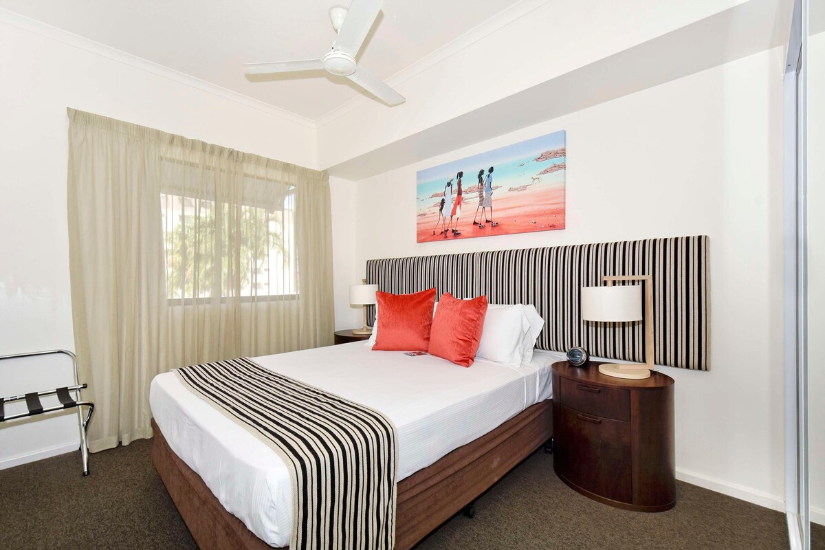 A hotel room with a bed, a nightstand, a lamp, a ceiling fan, a window with curtains, and a painting on the wall.