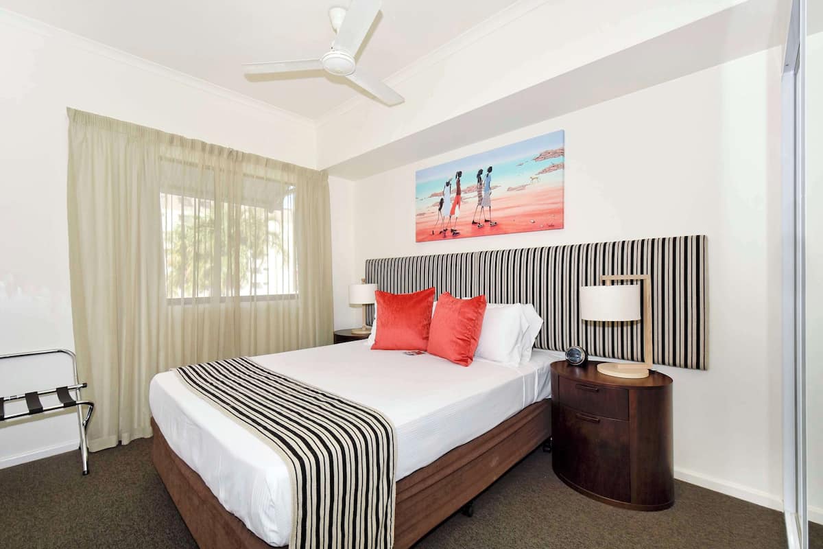 A hotel room with a large bed, a ceiling fan, a nightstand with a lamp, a painting on the wall, and a window with curtains.