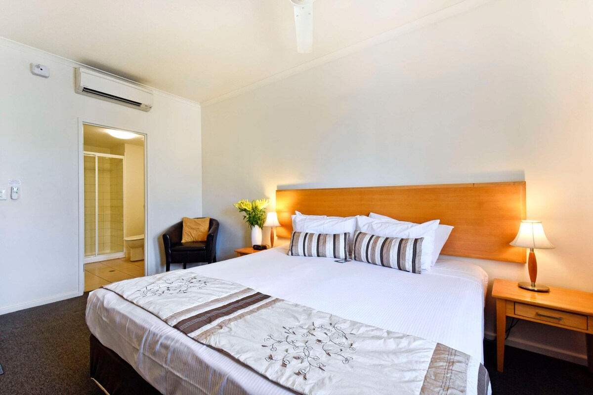 A hotel room with a large bed, bedside tables, a nightstand, a chair, a lamp, and an air conditioning unit.