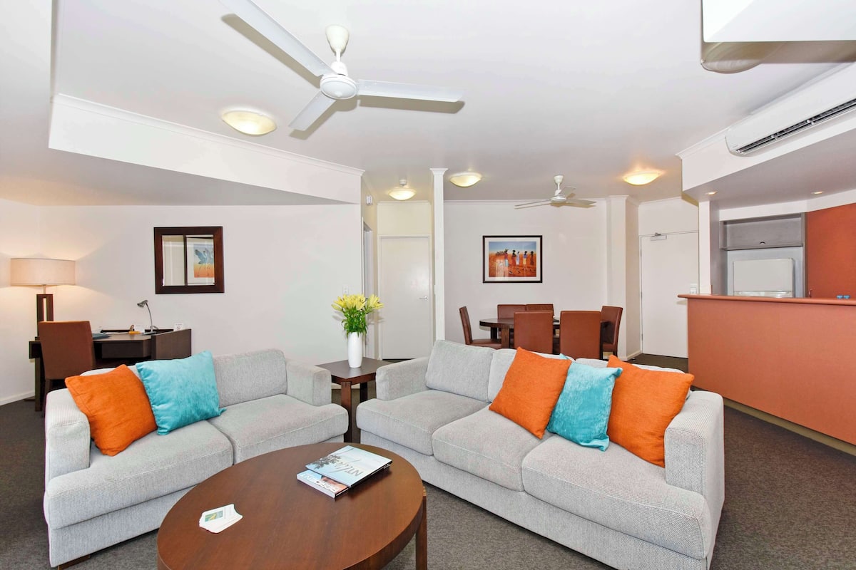 A modern living room with two grey sofas, a wooden coffee table, and a ceiling fan. There is a dining area with a table and chairs, and a reception desk.