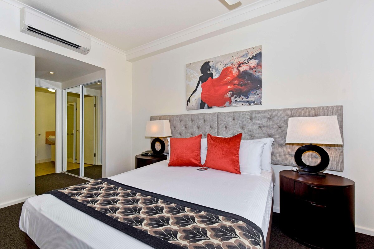 A hotel room with a bed, two bedside tables, a headboard, a painting, and a door leading to a bathroom.