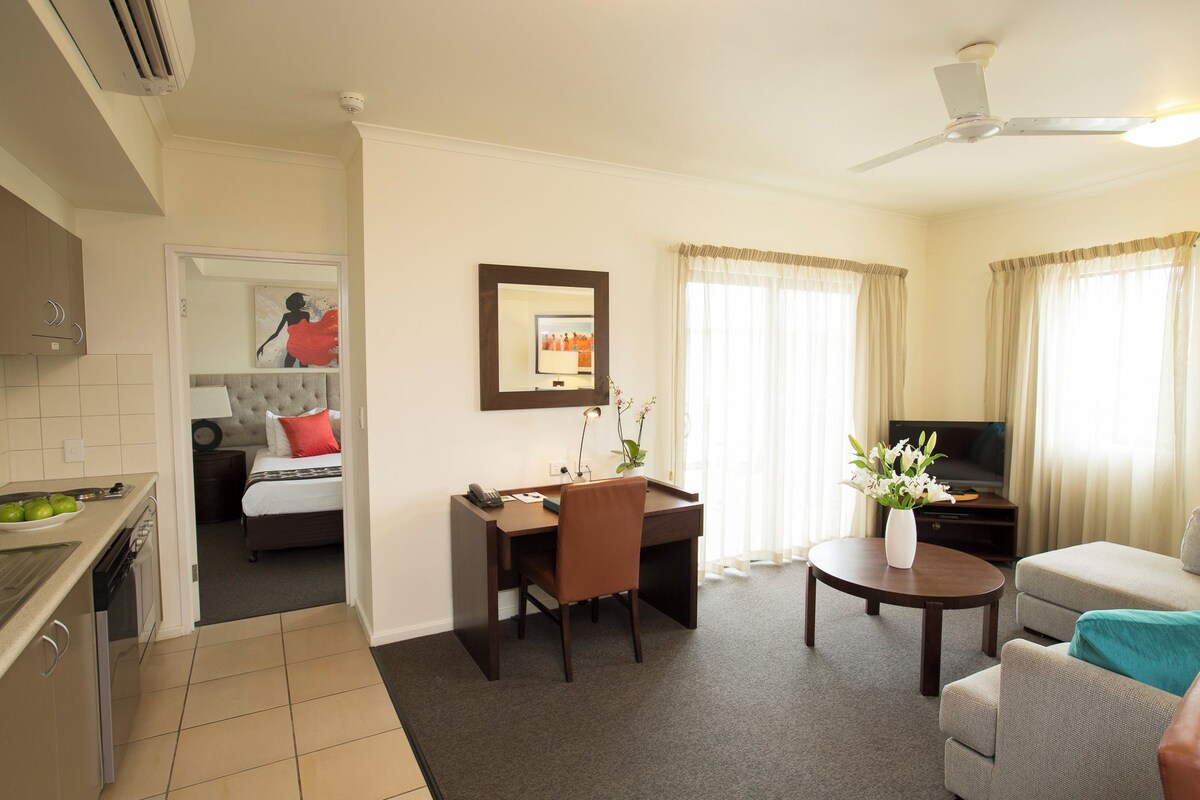 A hotel room with a kitchenette, a bed with a red pillow, a desk with a chair, a TV, and a sofa.