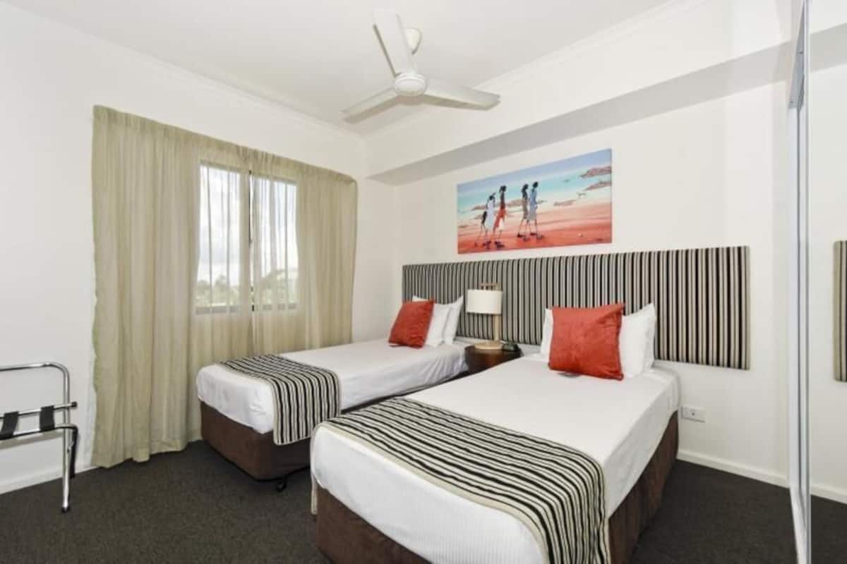 A hotel room with two beds, a ceiling fan, a window with curtains, a painting on the wall, and a bedside table with a lamp.