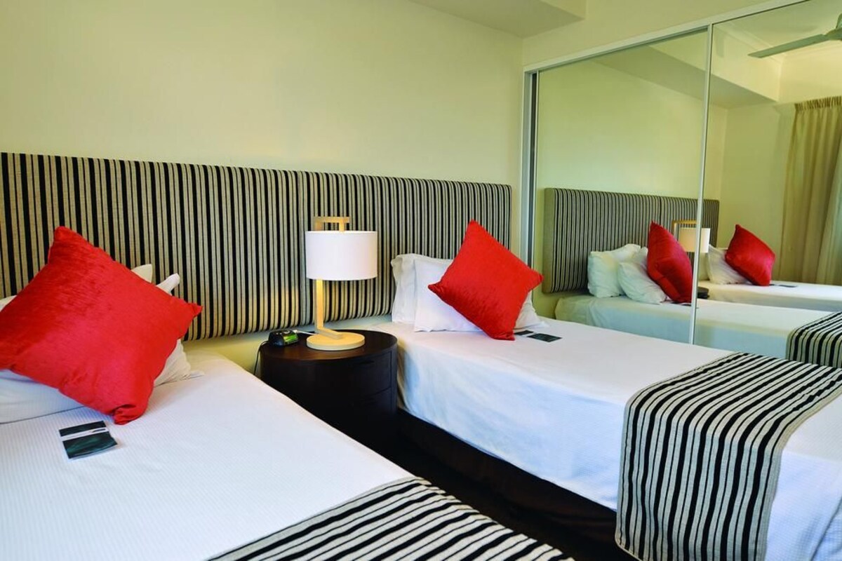 A hotel room with two beds, striped headboards, red pillows, a bedside table with a lamp, and a large mirror.