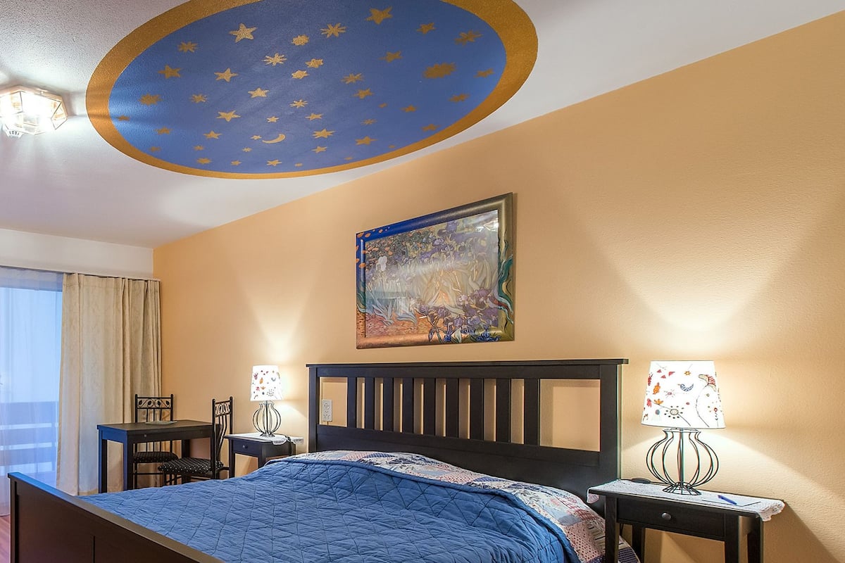 A hotel room with a bed, a chair, a table, and a painting on the wall.