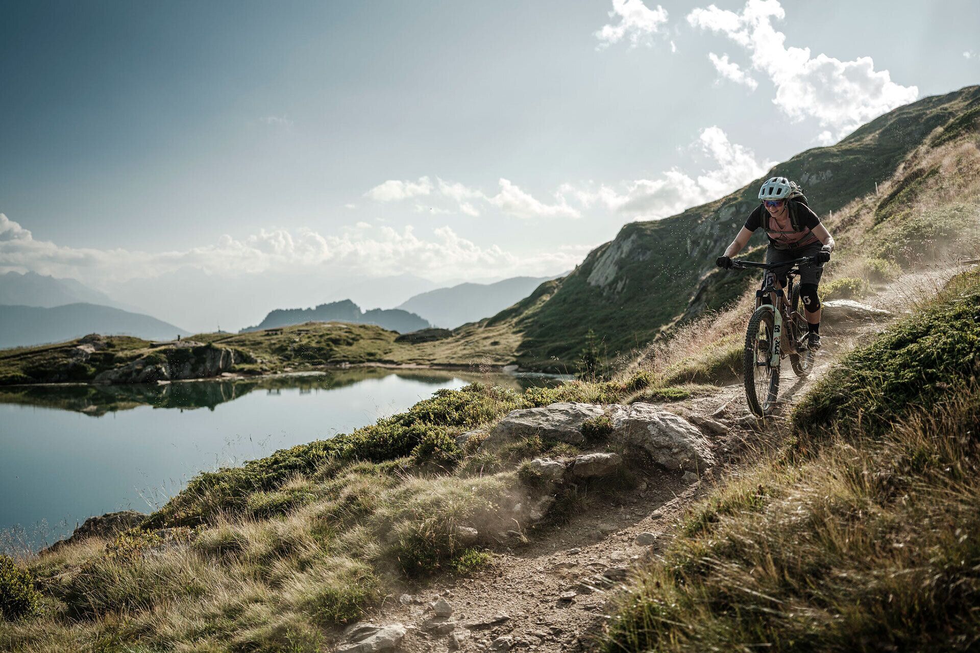 A mountain biker on a dirt trail with a lake and mountains in the background.