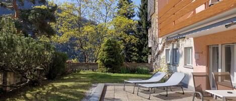 Eine Terrasse mit zwei Liegestühlen und einem Tisch, umgeben von einem Garten mit verschiedenen Bäumen und Sträuchern.