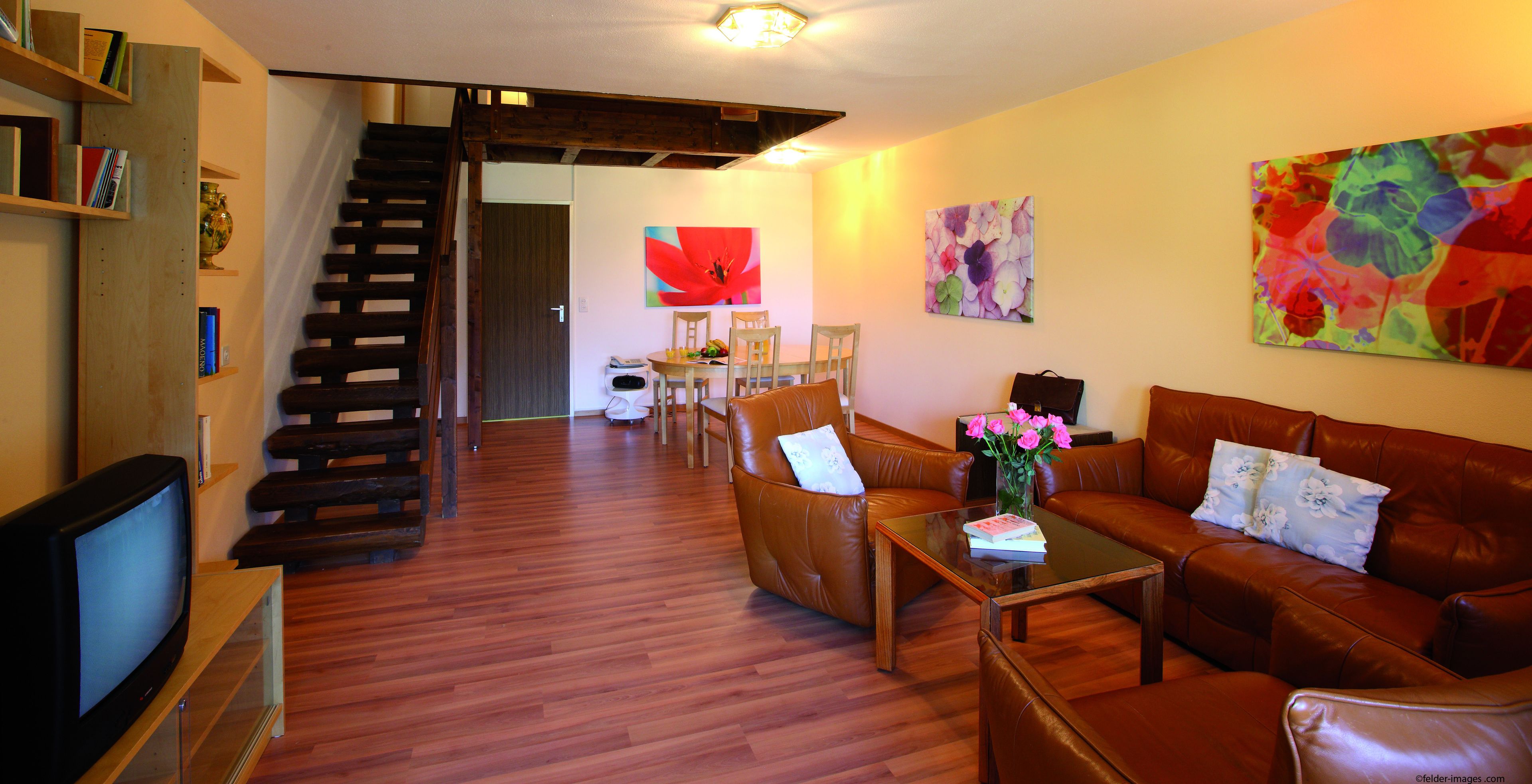 A living room with a wooden staircase, a brown leather sofa, a glass coffee table, and a TV on a wooden stand.