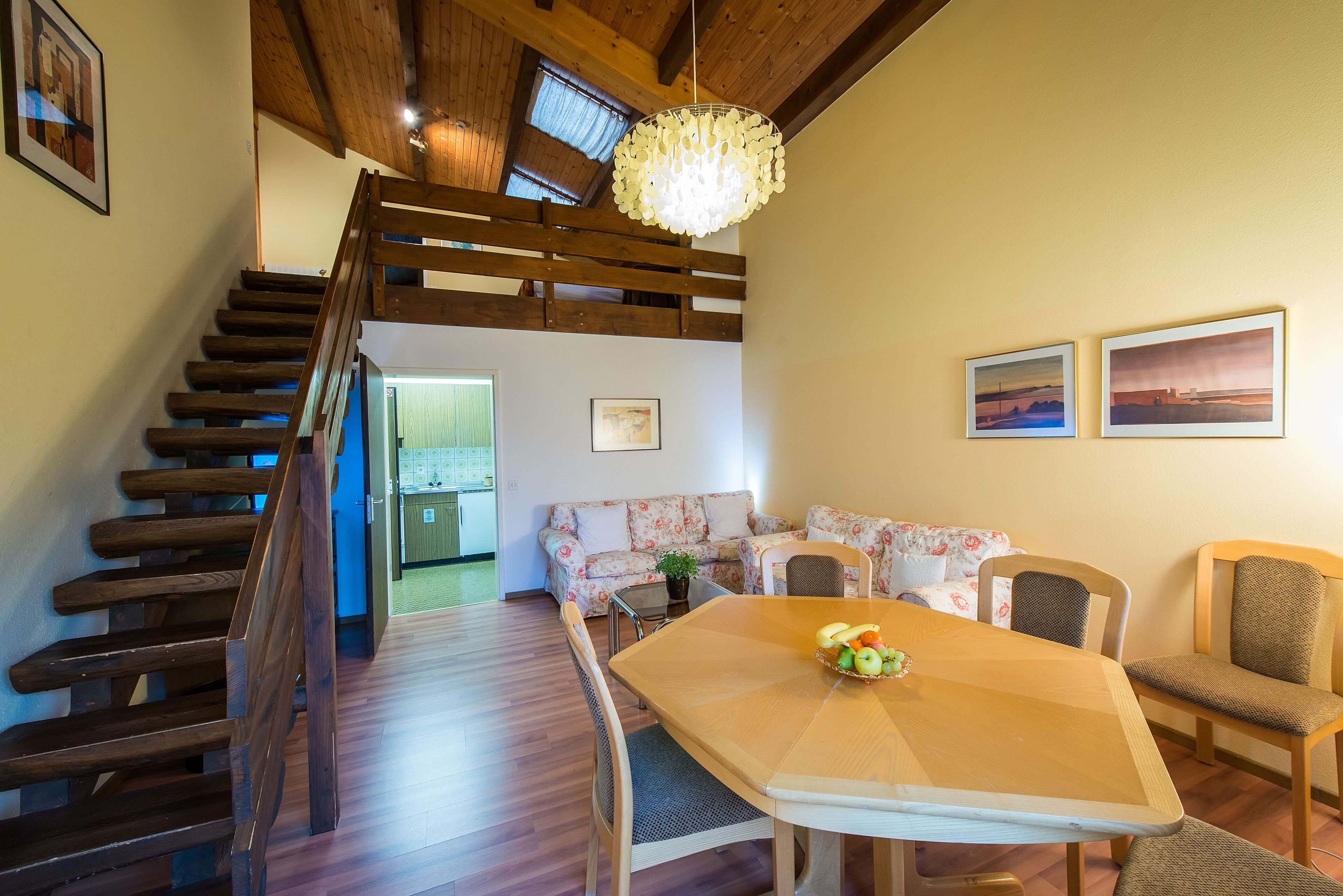 Una escalera de madera lleva a un espacio entrepiso con una araña, una mesa de comedor con fruta, un sofá floral y dos cuadros en la pared.