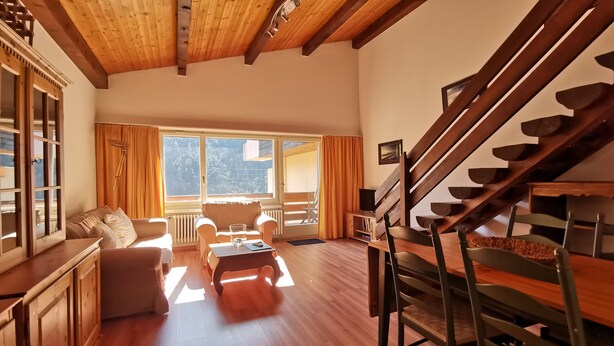 Een ruime woonkamer met een houten trap, een zithoek, een bijzettafeltje en uitzicht op de omgeving.