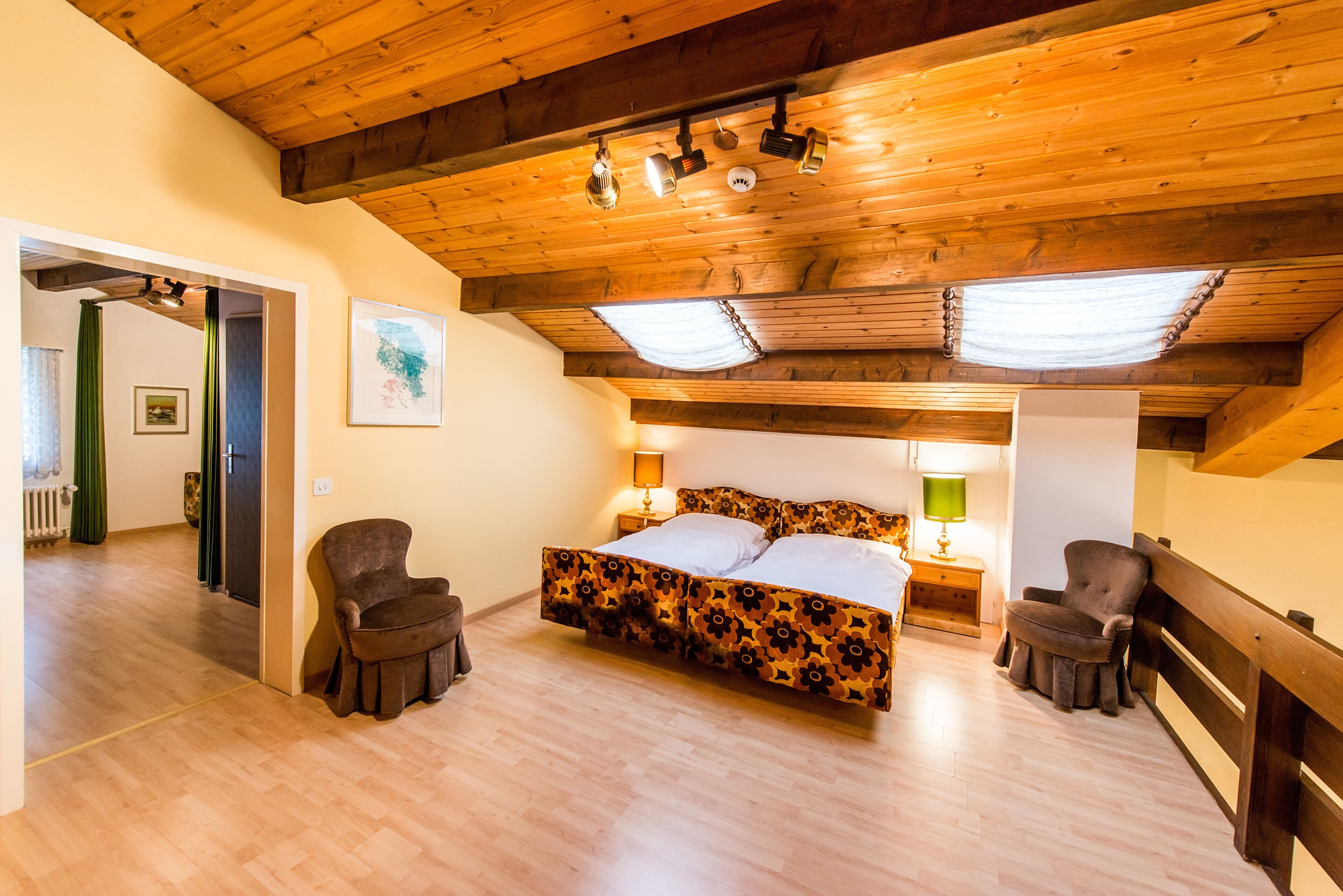 Een slaapkamer met een houten plafond, een bed met een bloemenbedekking, twee stoelen, een nachtkastje met een lamp en een schilderij aan de muur.