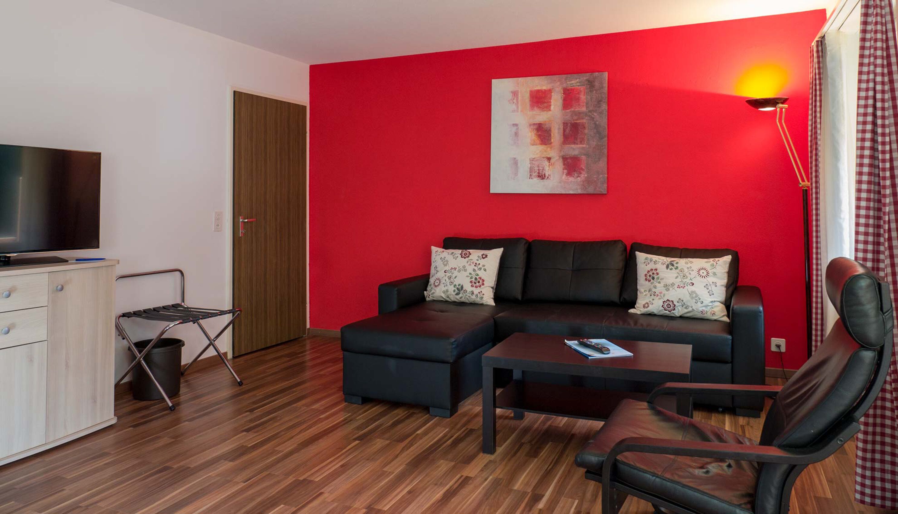 Un salon moderne avec un mur d’accent rouge, un canapé en cuir noir, une table basse en bois et un téléviseur posé sur un meuble.