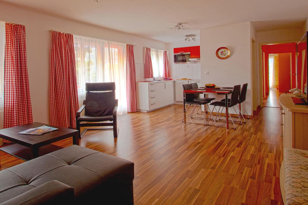 A modern living room with a sofa, a dining area with chairs and a table, a kitchen with white cabinets, and a red door.