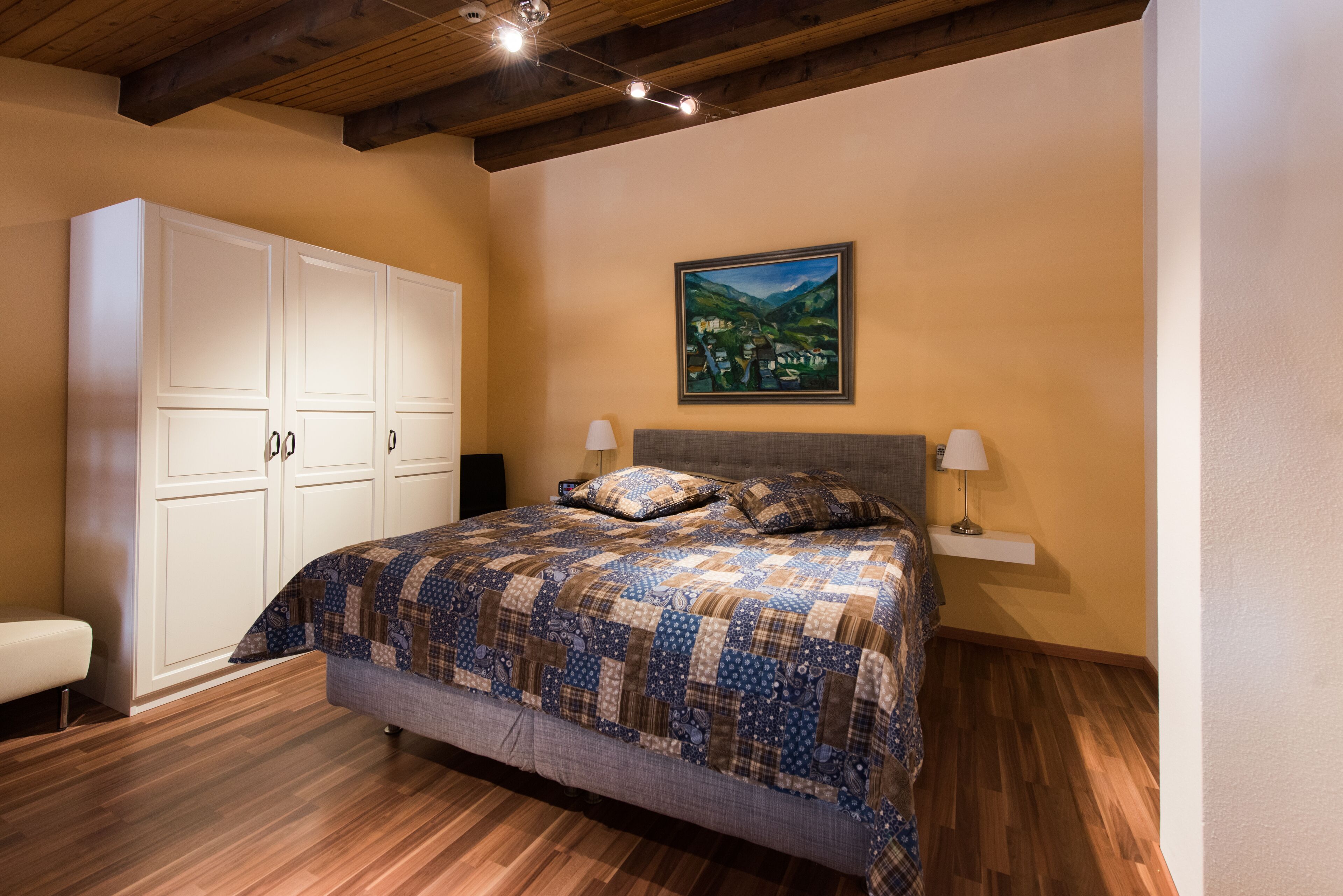 A bedroom with a large bed, wooden flooring, a white wardrobe, and a painting on the wall.