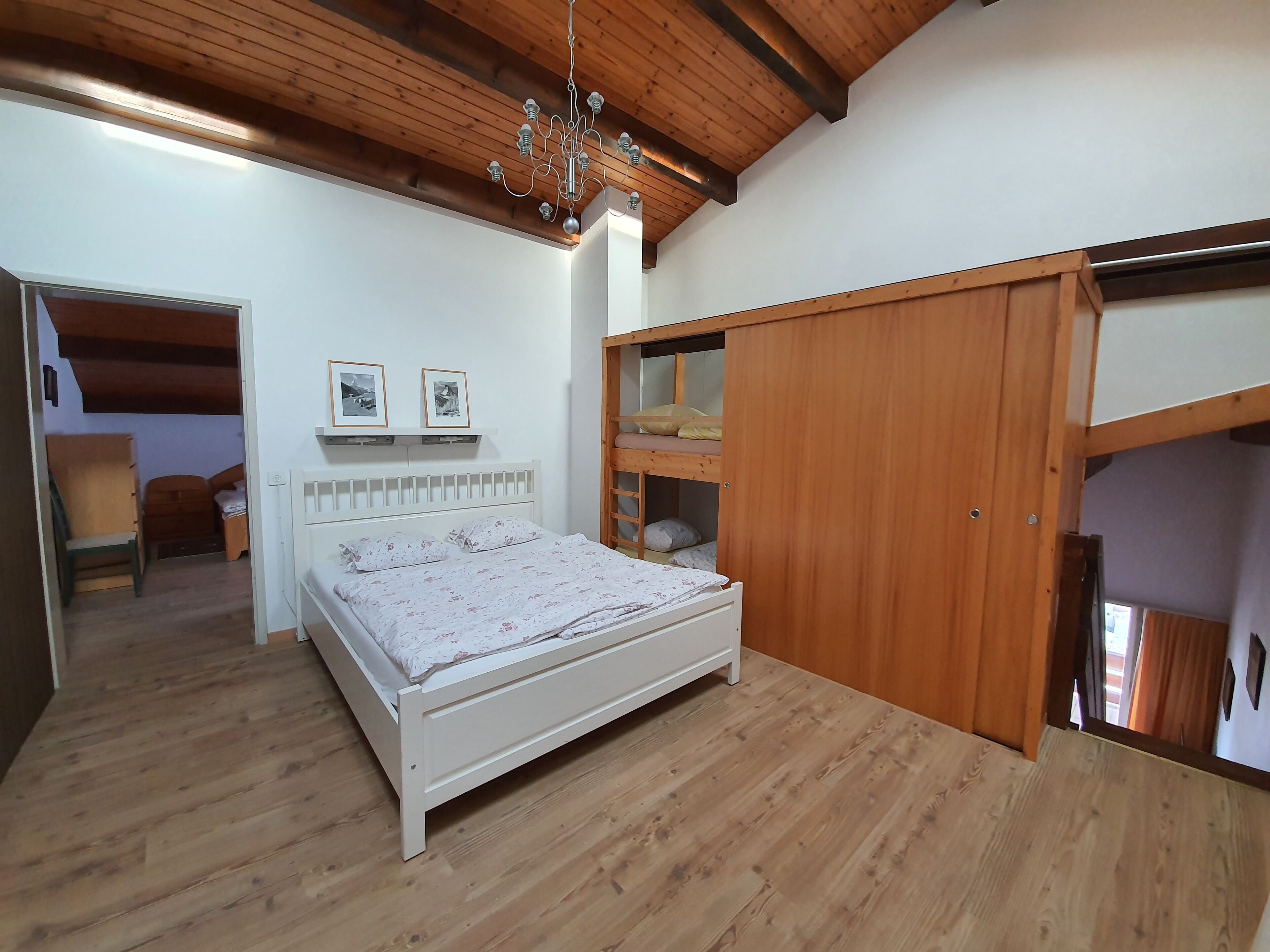 Een slaapkamer met een wit bed, houten vloer en een houten plafond. Er hangen twee ingelijste foto's aan de muur en staat een houten garderobe.