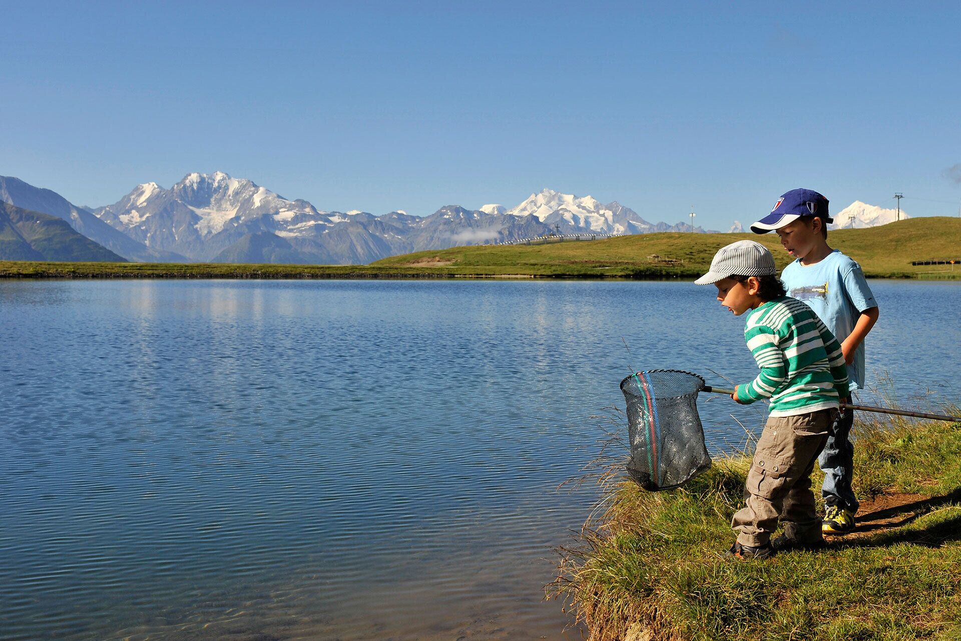Two children fishing by a calm lake with mountains in the background.