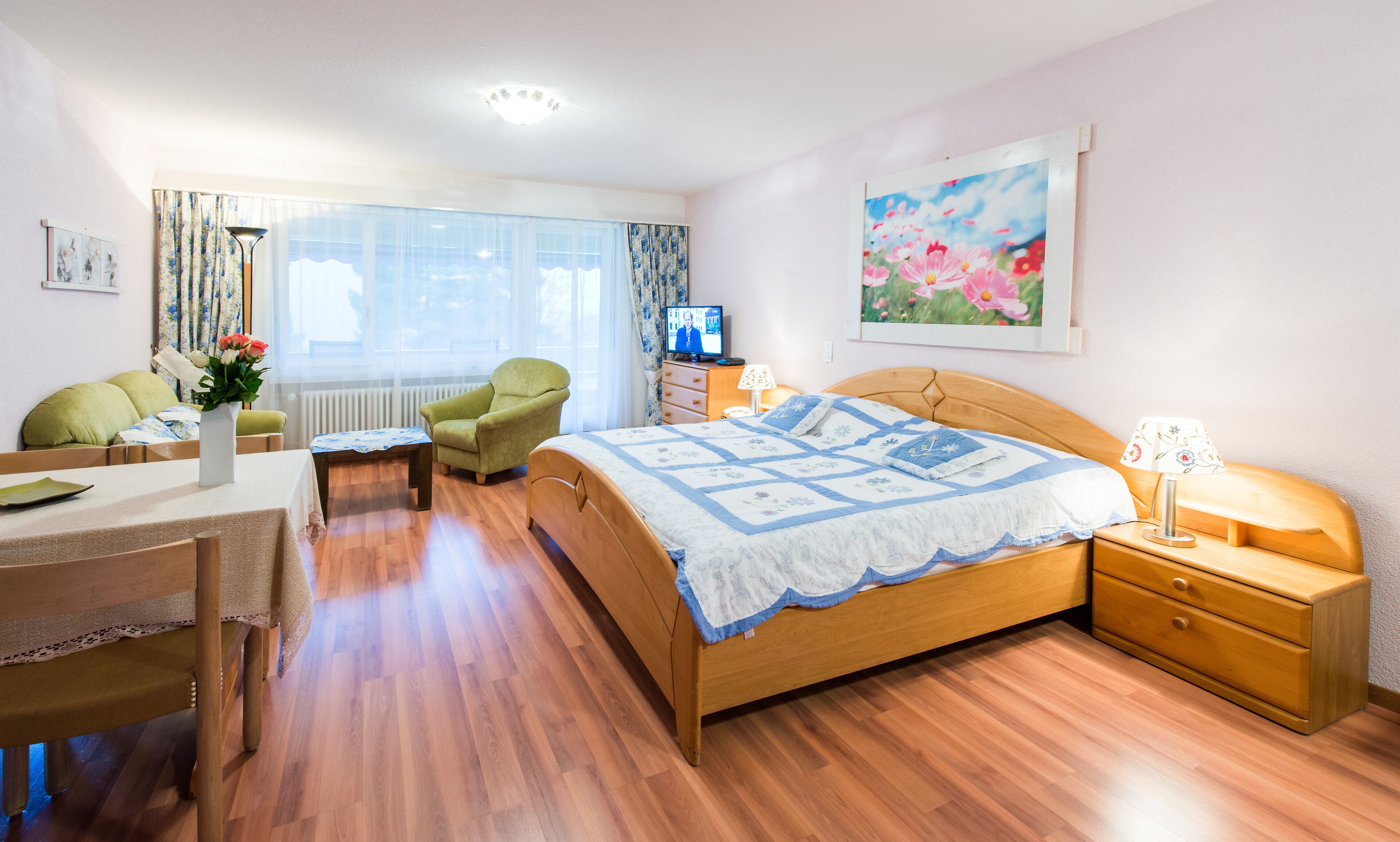 Un dormitorio bien iluminado con una cama de madera, un sillón verde, una mesita con un florero y un televisor sobre un aparador.