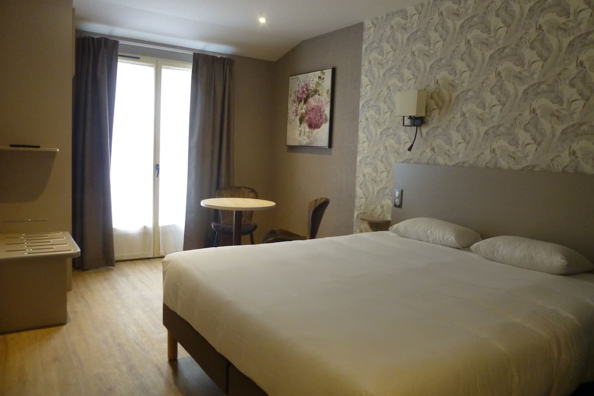 A hotel room with a large bed, a small round table, a chair, a desk, and a window with curtains.