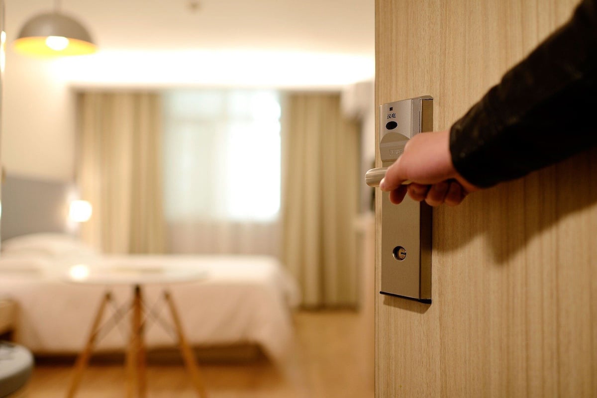 A hand turning a door handle in a hotel room with a bed and a lamp in the background.