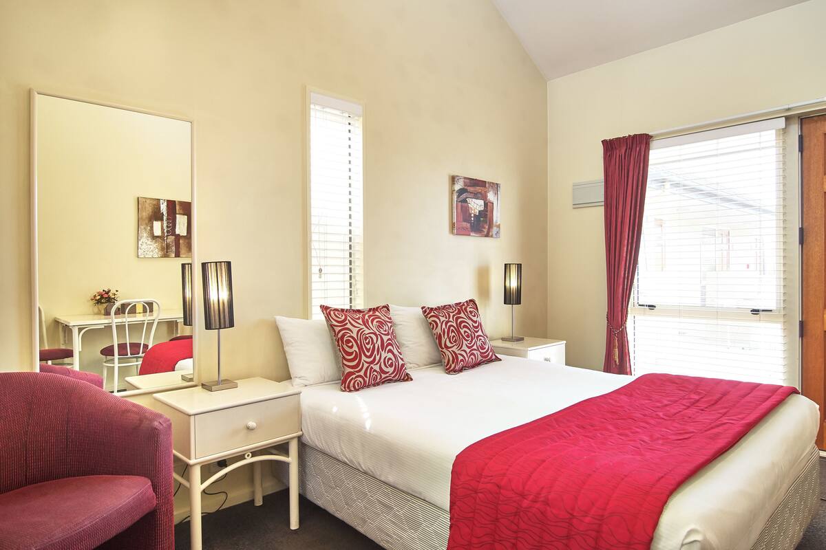 Une chambre d’hôtel avec un lit, un bureau, une chaise, un miroir et une fenêtre avec des rideaux.