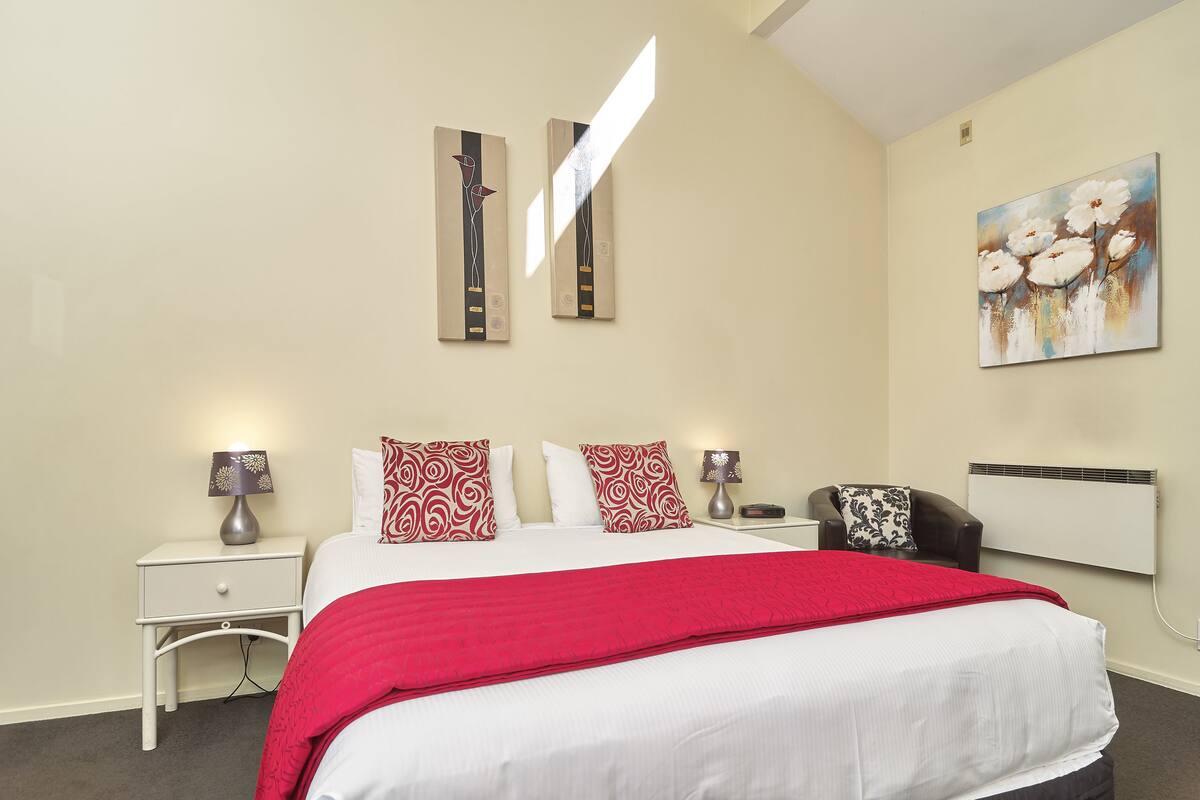 Une chambre d’hôtel moderne avec un grand lit, un plaid rouge, deux tables de chevet avec des lampes, une chaise et un tableau au mur.