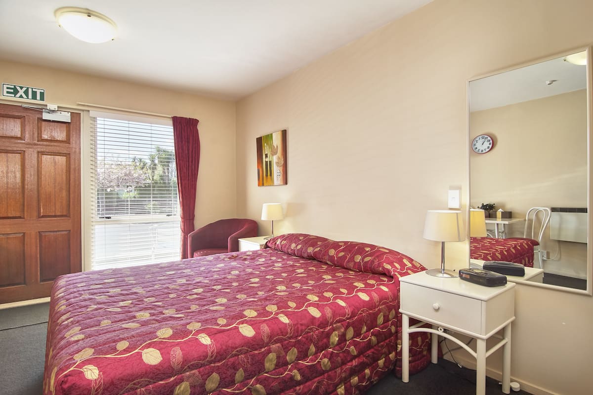 Une chambre d’hôtel avec un lit, une table de chevet, une lampe, un miroir, une horloge et un panneau indiquant la sortie.