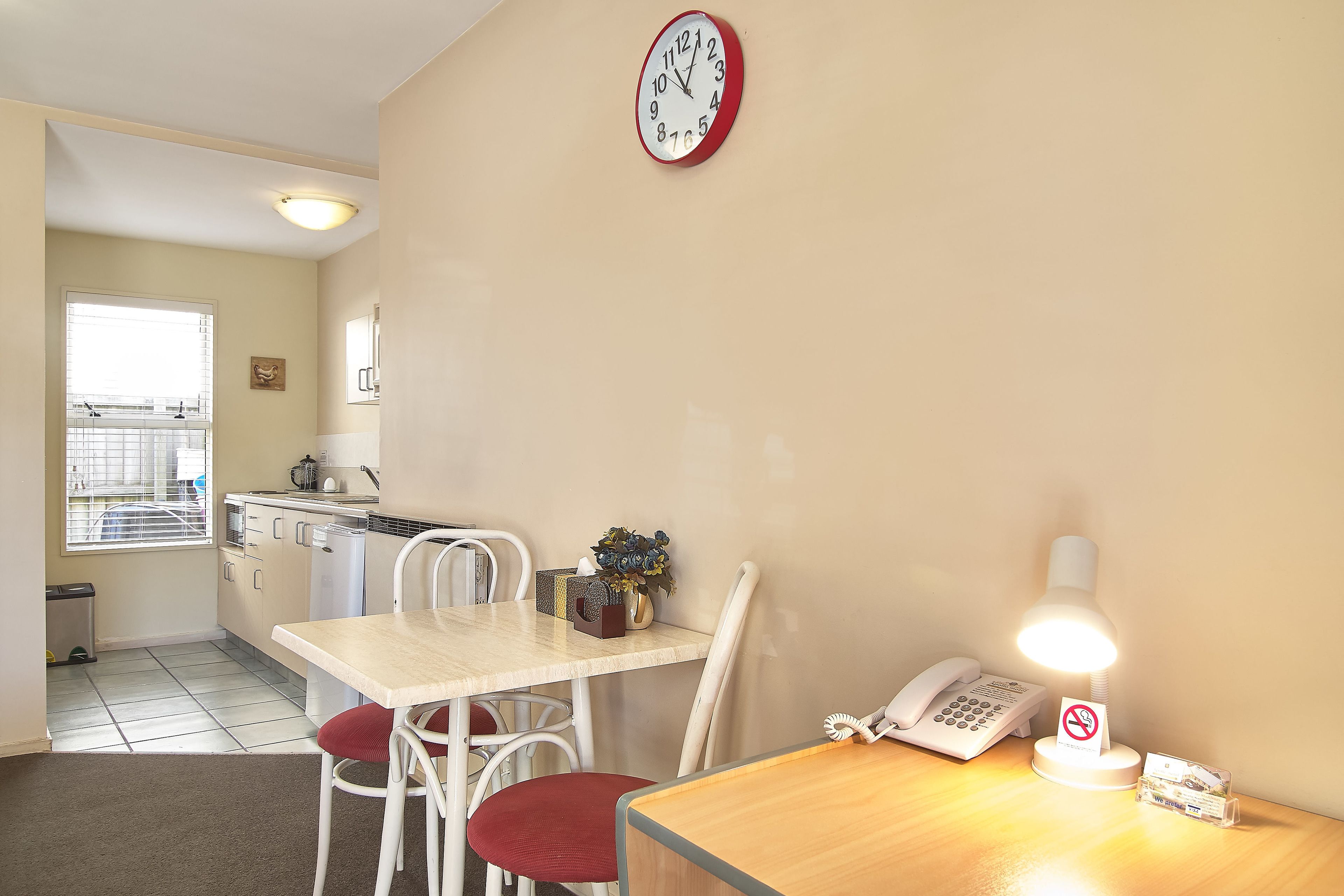 A small, well-lit room with a wooden desk, a chair, a clock, a lamp, and a phone.