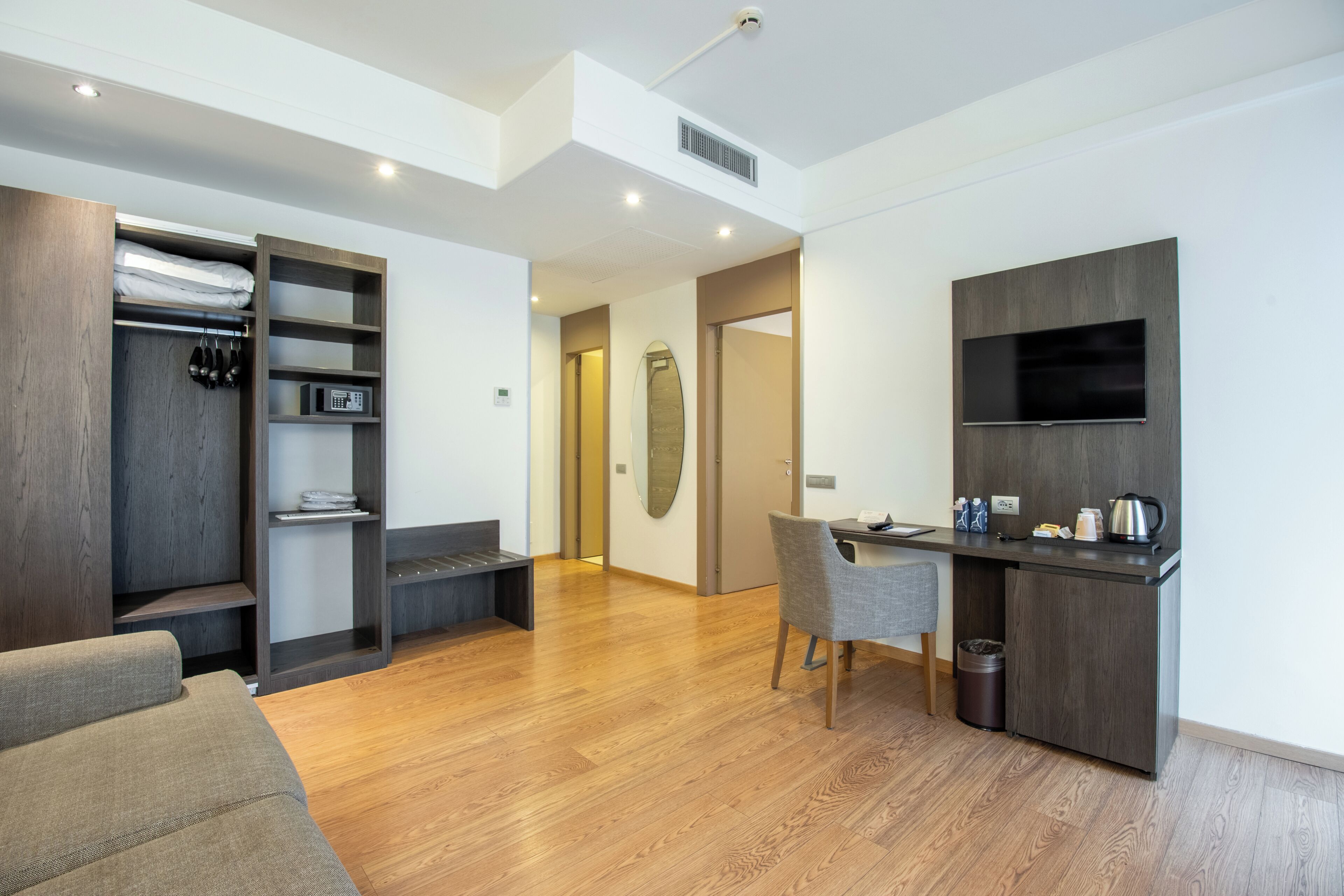 A modern hotel room with a wooden floor, a sofa, a desk with a chair, a TV, a coat rack, and a wardrobe.