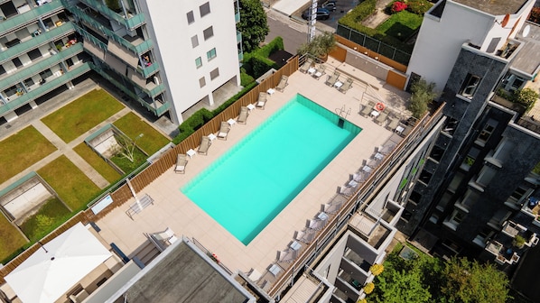 A rooftop pool with sun loungers and a wooden fence, surrounded by high-rise buildings.