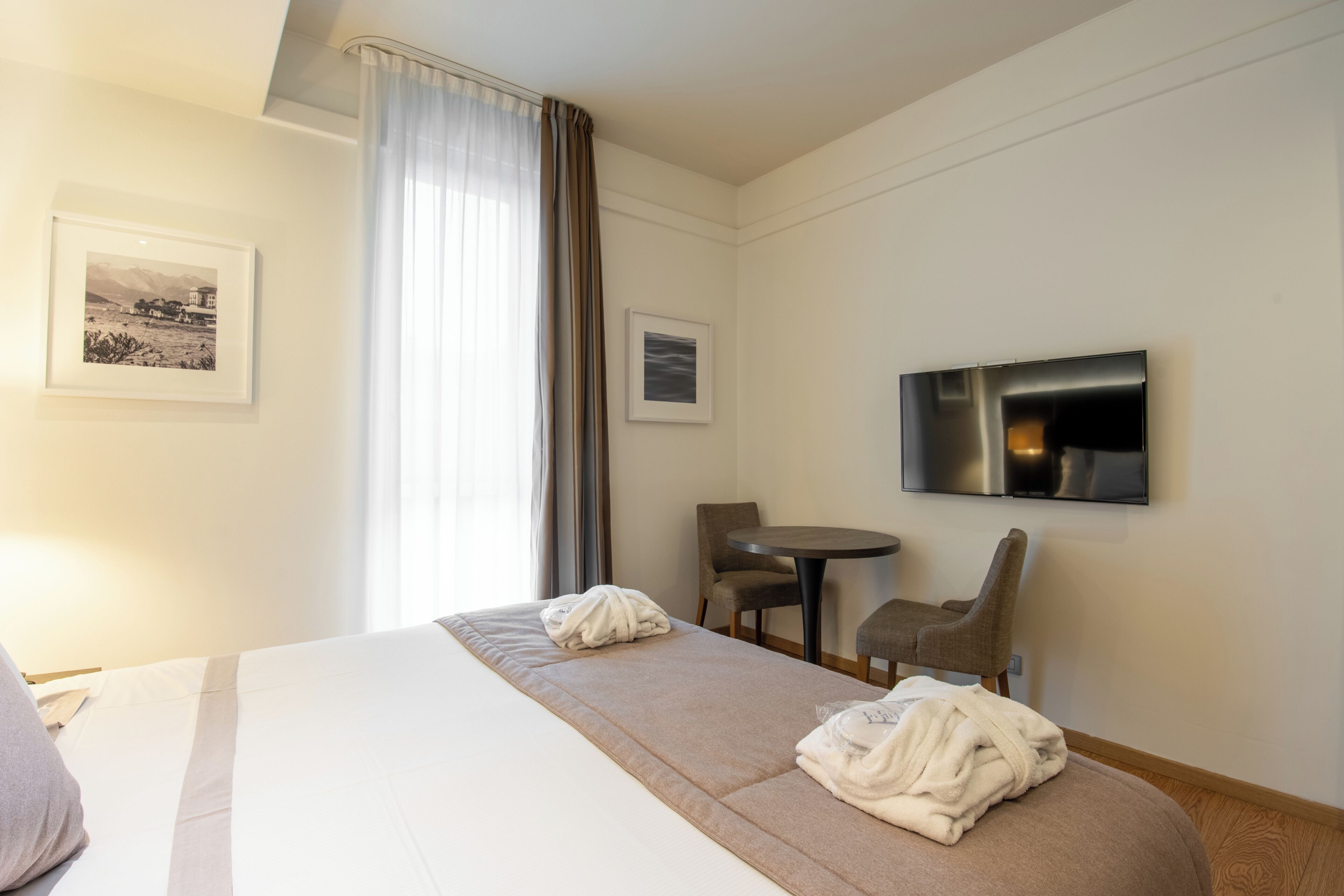 A hotel room with a bed, two chairs, a small table, a TV, and two framed pictures on the wall.
