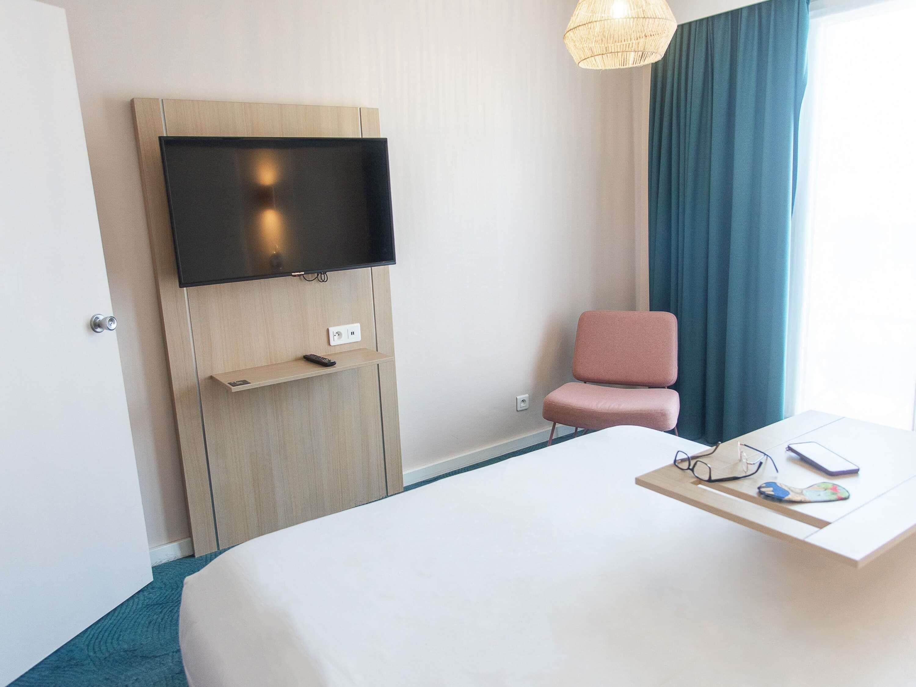 A hotel room with a bed, a TV mounted on a wooden panel, a pink chair, a desk with glasses and a phone, and a window with curtains.