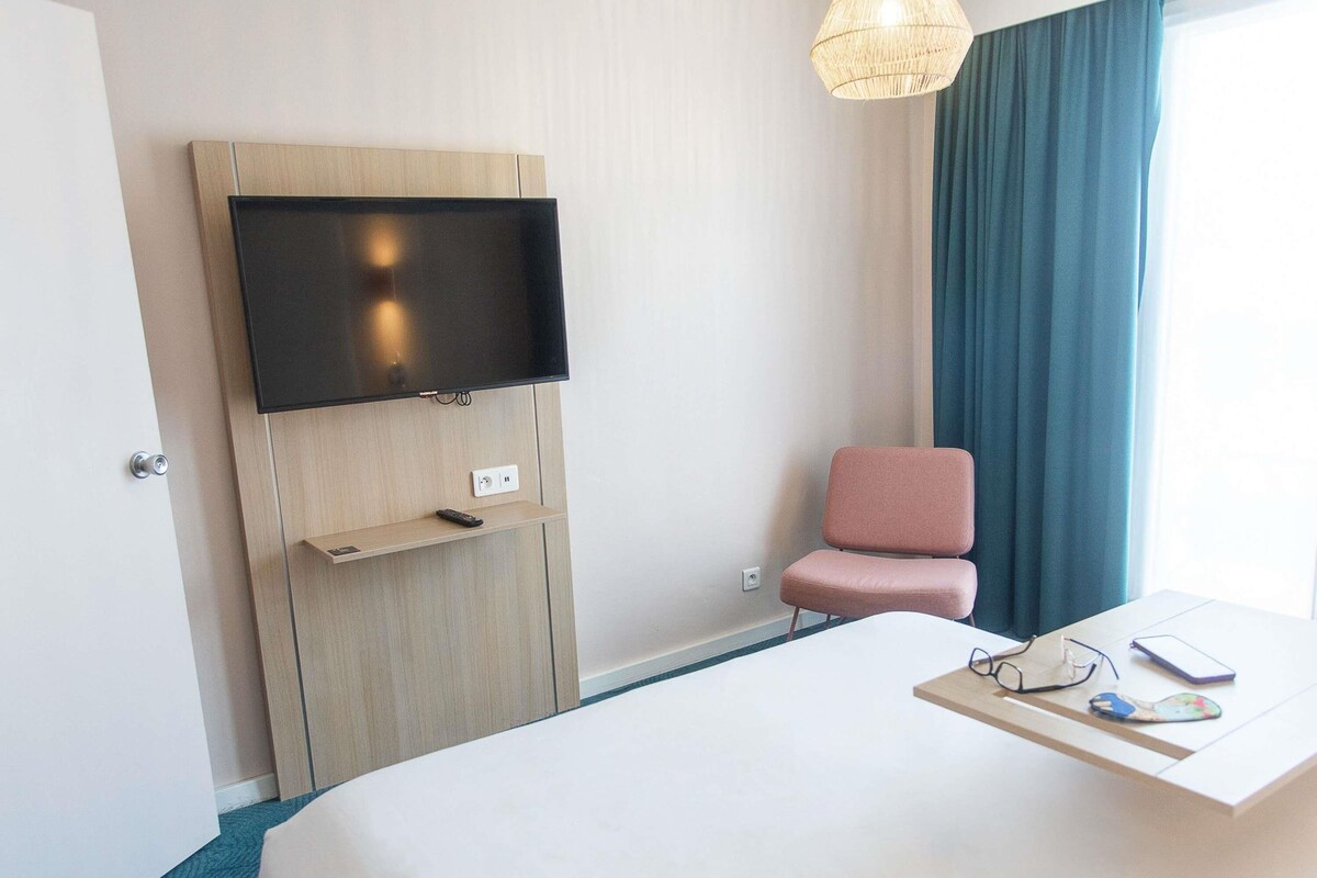 A hotel room with a bed, a TV mounted on a wooden panel, a pink chair, a desk with glasses and a phone, and a window with curtains.